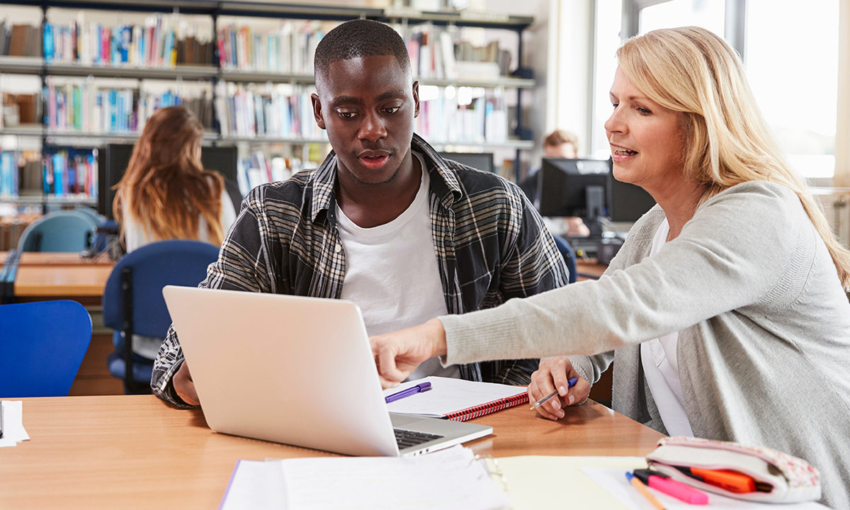 A teenage boy sits with a teacher in the library and they work together on his laptop